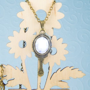 May include: A gold-tone necklace with a vintage-style hand mirror pendant. The mirror has a clear glass surface and a decorative frame. The necklace is displayed on a wooden stand with a floral design.