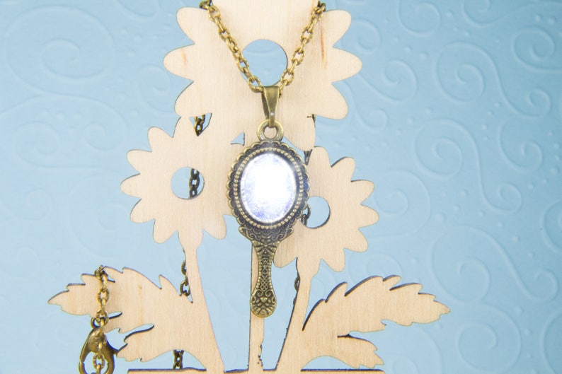 May include: A gold-tone necklace with a vintage-style hand mirror pendant. The mirror has a clear glass surface and a decorative frame. The necklace is displayed on a wooden stand with a floral design.