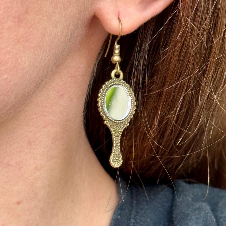 May include: Gold-toned dangle earrings featuring a small, round mirror with a decorative frame and a handle in the shape of a stylized hand.