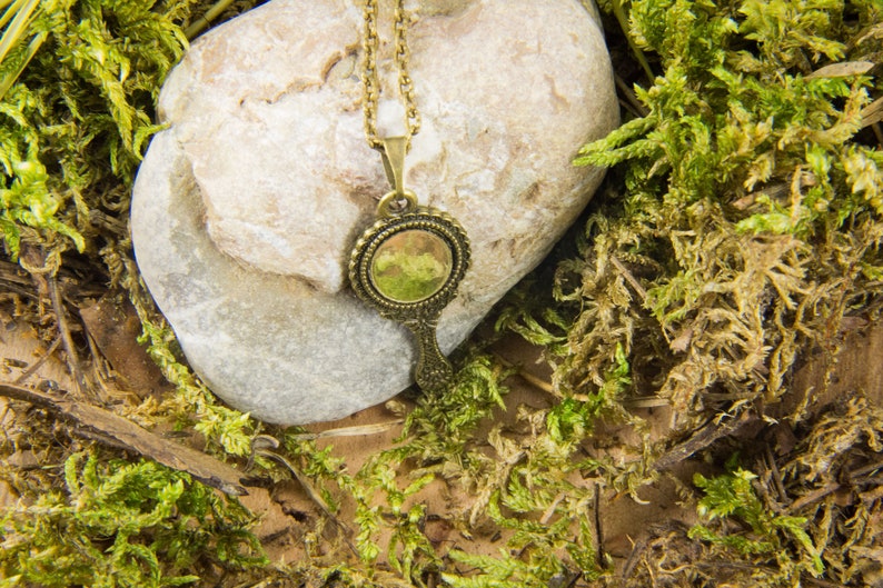 May include: A gold-toned necklace with a round, ornate mirror pendant. The pendant has a green glass mirror and is surrounded by a decorative frame. The necklace is lying on a rock and moss.