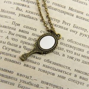 May include: A gold-toned necklace with a small, round, ornate mirror pendant. The mirror is surrounded by a decorative frame and the necklace is made of a chain with a delicate design. The necklace is lying on a page of a book with text.