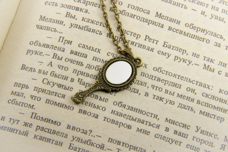 May include: A gold-toned necklace with a small, round, ornate mirror pendant. The mirror is surrounded by a decorative frame and the necklace is made of a chain with a delicate design. The necklace is lying on a page of a book with text.