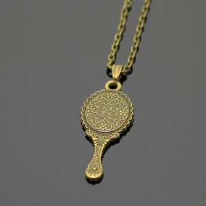 May include: A gold-toned necklace with a pendant shaped like a hand mirror. The mirror has an ornate design with a floral pattern.