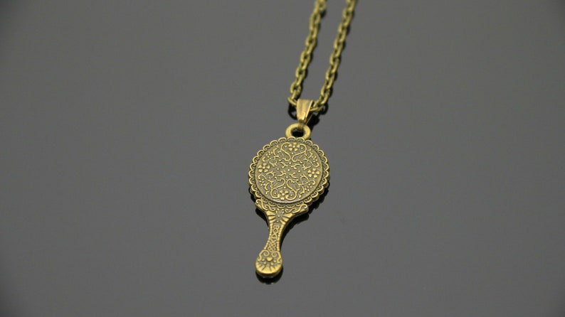 May include: A gold-toned necklace with a pendant shaped like a hand mirror. The mirror has an ornate design with a floral pattern.
