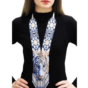 May include: A beaded necklace featuring a detailed tiger face design in shades of blue, white, and tan. The necklace has a long, vertical design with a fringe at the bottom. The background is a solid black.