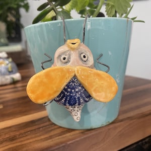 May include: A whimsical ceramic bee ornament with orange wings and a blue and white patterned body, attached to a light blue plant pot. The bee has a surprised expression and wire antennae. The pot contains green plants.