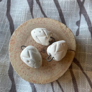 May include: Three white ceramic beads with a textured surface and a small metal loop. The beads are arranged on a wooden surface.