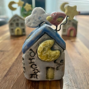 May include: A ceramic house figurine with a blue roof, a yellow moon, and a cloud. The word "house" is written on the front. A red and yellow bird sits on top of the house. Other similar figurines are in the background.