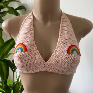 May include: A light pink crochet halter top with rainbow patches. The top is handmade with a textured stitch and features a V-neck design. The rainbow patches are in a classic rainbow colour pattern.