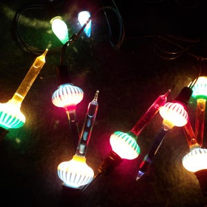 May include: A string of vintage Christmas lights with colorful bulbs. The bulbs have a ribbed, globe-like design with green, red, and white accents. The lights are strung on a dark wire, creating a festive and nostalgic look.