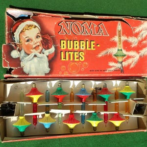 May include: A vintage Noma Bubble-Lites Christmas light set in a red cardboard box featuring a child in a Santa hat. The box contains multiple colorful glass bubble light bulbs with plastic bases in red, yellow, green, and blue colors.