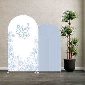 Puede incluir: Dos paneles de fondo arqueados. Uno es blanco con ilustraciones florales azules y el texto "Bride to Be". El otro panel es azul claro con un diseño de líneas verticales curvas. Ambos están sobre soportes plateados.