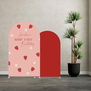 May include: Two arched backdrop panels, one light pink with a strawberry and daisy pattern and the text "Amelia's BERRY FIRST Birthday" in red, and one solid dark red panel. The panels are freestanding on metal bases with a thin frame design.