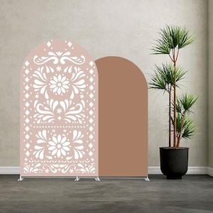 May include: Two arched backdrop panels. One is a light pink with a white floral and geometric design. The other is a solid brown. The panels are on silver stands. A tall potted plant is in the background.