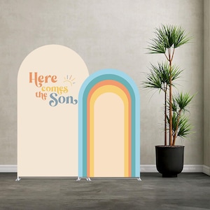 May include: Two arched backdrop banners. One is cream-coloured with the text "Here comes the Son" in orange and blue. The other banner features a rainbow design in shades of blue, orange, and yellow. A potted plant is to the right.