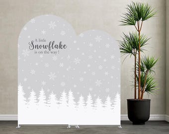 May include: A dual-arched backdrop display featuring a light grey background with white snowflake patterns and a white silhouette of a pine forest at the base. The text "A little Snowflake is on the way!" is printed in dark grey script.