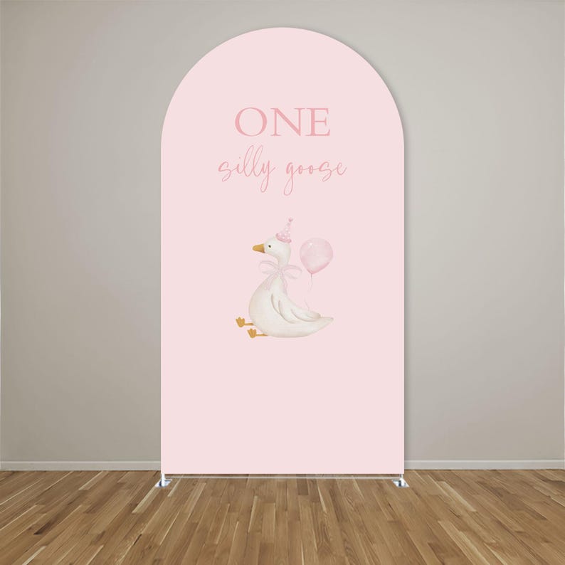 May include: A pink arch-shaped backdrop with the words "ONE silly goose" in light pink. A watercolor illustration of a white goose wearing a party hat and holding a pink balloon is featured below the text. The backdrop is supported by a silver metal frame.