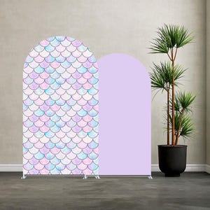 Op de afbeelding: Twee gebogen achtergrondstandaards. Eén heeft een zeemeerminstaartpatroon in pastelblauw, paars en roze. De andere is effen lavendel. Beide staan op zilveren standaards. Rechts staat een hoge plant in een pot.