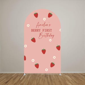 May include: An arched backdrop cover in a soft pink hue featuring a repeating pattern of red strawberries and small white daisies. The center text reads Amelia's BERRY FIRST Birthday in a combination of script and block lettering in red.