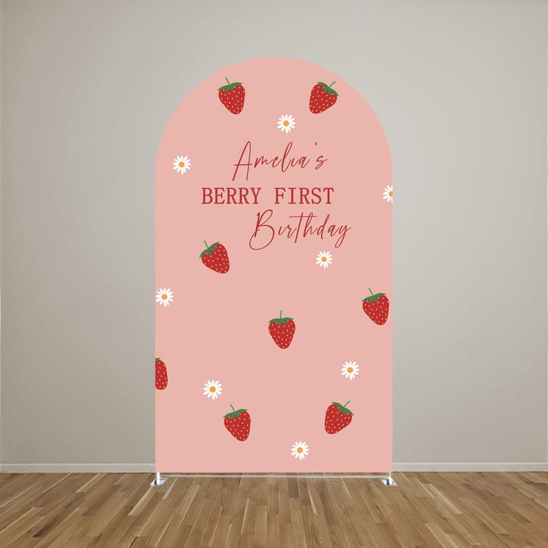 May include: An arched backdrop cover in a soft pink hue featuring a repeating pattern of red strawberries and small white daisies. The center text reads Amelia's BERRY FIRST Birthday in a combination of script and block lettering in red.