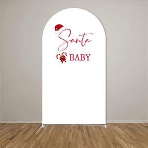 May include: A white arched backdrop cover featuring a red Santa hat graphic above the red cursive text Santa and the block text BABY, accompanied by a red and white candy cane illustration with a green holly accent on the left side.
