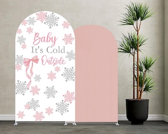 May include: Two arched backdrop stands. One has the text "Baby It's Cold Outside" with snowflakes and a pink bow. The other is a solid pink colour with vertical lines. A potted plant is in the background.