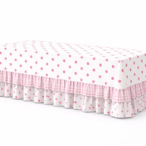 Puede incluir: Una cubierta de mesa rectangular con base blanca y lunares rosas. Los bordes presentan un patrón de cuadros rosa y blanco, complementado con un ribete con volantes. El diseño general es suave y femenino.