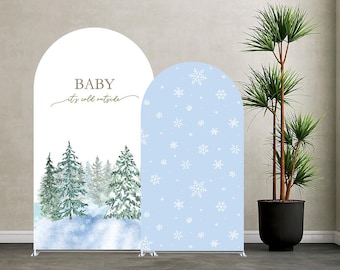 May include: Two arched backdrop covers featuring winter themes. The left cover is white with the text "BABY it's cold outside" above a watercolour forest of green pine trees and snow. The right cover is light blue with a white snowflake pattern.