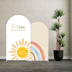 May include: Two arched backdrop banners. One white banner features a watercolour sun and the text "First Trip Around The Sun." The other banner is beige with a watercolour rainbow. A potted plant is to the right.