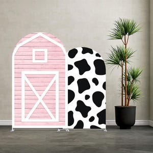 Könnte beinhalten: Zwei bogenförmige Hintergrundständer. Einer ist ein rosa Scheunendesign mit einer weißen Tür und einem Fenster. Der andere hat einen schwarz-weißen Kuhdruck. Rechts ist eine hohe Topfpflanze.
