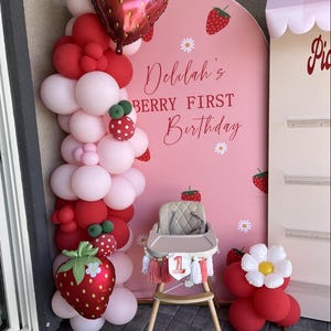 May include: A pink arched backdrop with strawberry and daisy graphics, featuring the text Delilah's BERRY FIRST Birthday. A balloon garland in red and light pink with strawberry foil balloons, a beige high chair, and a red balloon cluster.