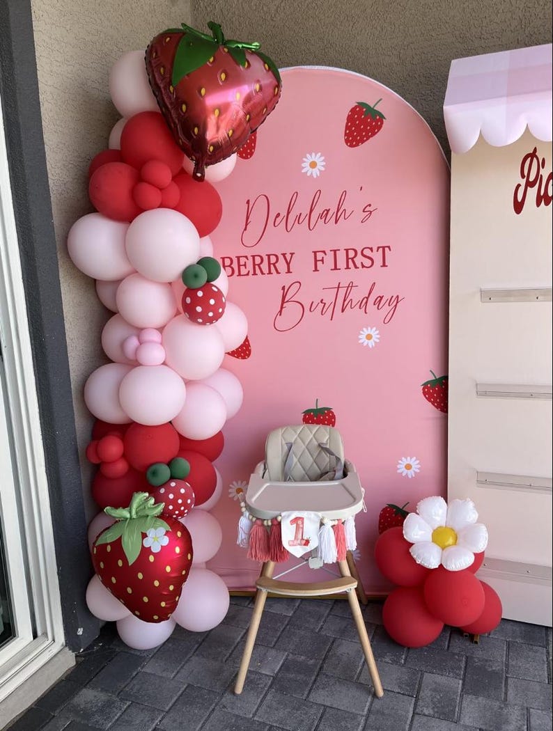 May include: A pink arched backdrop with strawberry and daisy graphics, featuring the text Delilah's BERRY FIRST Birthday. A balloon garland in red and light pink with strawberry foil balloons, a beige high chair, and a red balloon cluster.
