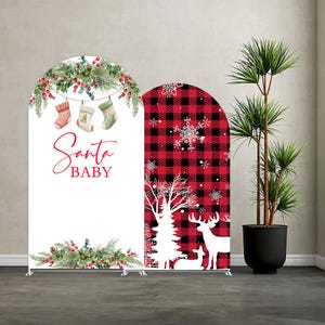 May include: Two arched backdrop covers featuring Christmas themes. The left cover is white with hanging stockings, pine branches, red berries, and the text Santa BABY. The right cover features a red and black buffalo plaid pattern with white deer silhouettes.