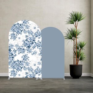 Könnte beinhalten: Zwei bogenförmige Hintergrundpaneele. Ein Paneel zeigt ein blaues Blumenmuster auf weißem Grund. Das andere Paneel ist einfarbig hellblau. Die Paneele werden von silbernen Metallständern getragen. Eine Topfpflanze befindet sich im Hintergrund.