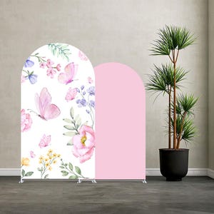 May include: Two arched backdrop stands. One features a white background with pink butterflies and floral designs. The other is solid pink. The stands are supported by silver metal frames. A potted plant is to the right.