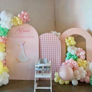 May include: A whimsical party scene with a high chair, balloons, and decorative arches. The backdrop features a pink arch with a goose illustration and text that reads "Atiana is one silly goose!" Pastel-colored balloons adorn the scene.