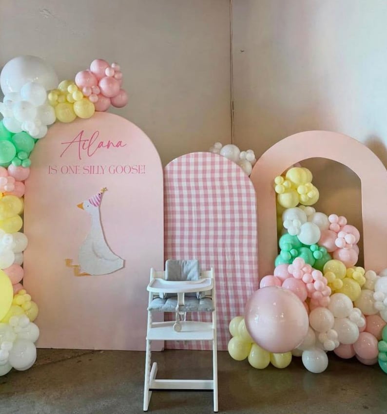 May include: A whimsical party scene with a high chair, balloons, and decorative arches. The backdrop features a pink arch with a goose illustration and text that reads "Atiana is one silly goose!" Pastel-colored balloons adorn the scene.