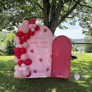 May include: A pink arched backdrop featuring strawberry and daisy illustrations with the text Veronica's BERRY FIRST Birthday, decorated with a cluster of red and light pink balloons, alongside a solid dark pink arched panel and a strawberry cutout.