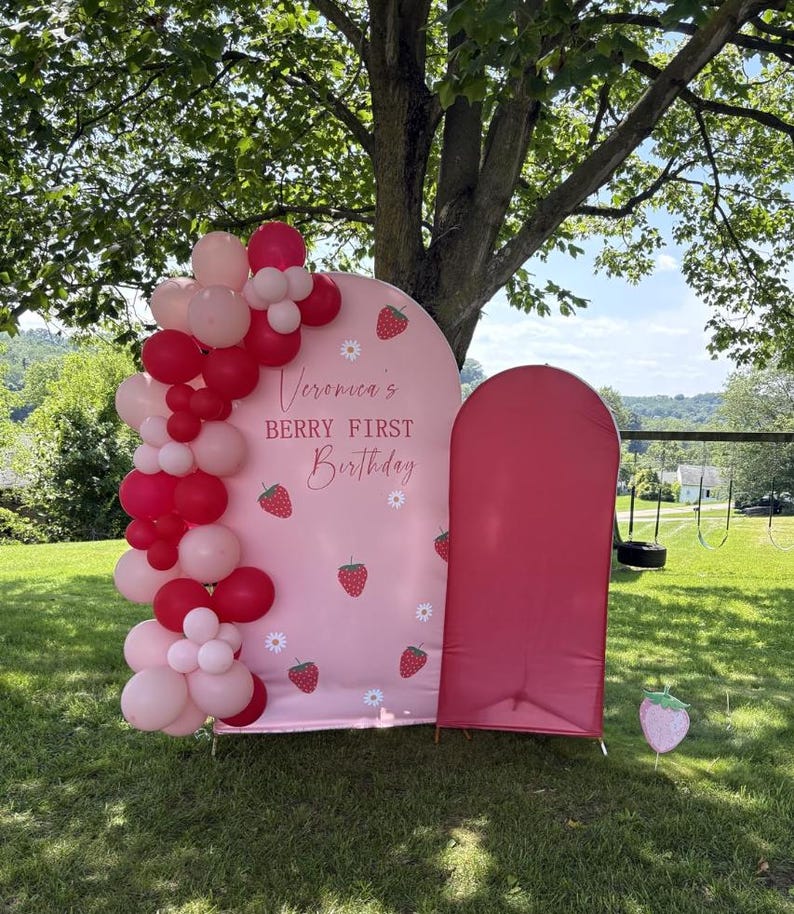 May include: A pink arched backdrop featuring strawberry and daisy illustrations with the text Veronica's BERRY FIRST Birthday, decorated with a cluster of red and light pink balloons, alongside a solid dark pink arched panel and a strawberry cutout.