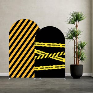 May include: Two arched backdrop displays. One has yellow diagonal stripes on a black background. The other has black background with yellow caution tape that reads "UNDER CONSTRUCTION". A potted plant is to the right.