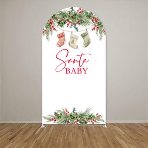 May include: An arched backdrop featuring a white background with a top and bottom border of green pine branches, red berries, and pinecones. Three hanging Christmas stockings are centred below the top border with the red text Santa BABY.
