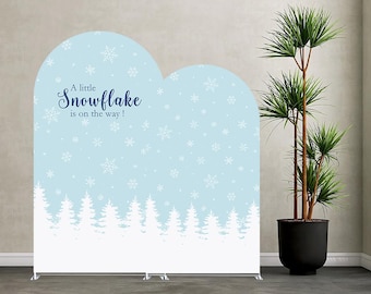 May include: A light blue arched backdrop featuring a white winter forest silhouette at the base and white snowflake patterns throughout. The text "A little Snowflake is on the way!" is printed in dark blue script on the upper left side of the panel.