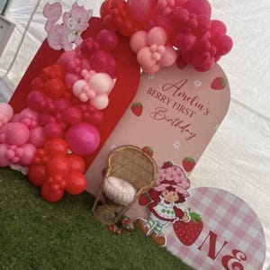 May include: A birthday display featuring a red and pink balloon arch, a pink arched backdrop with the text Amelia's BERRY FIRST Birthday, a wicker chair with a white cushion, a Strawberry Shortcake cutout, and a pink gingham sign with the letter ONE.