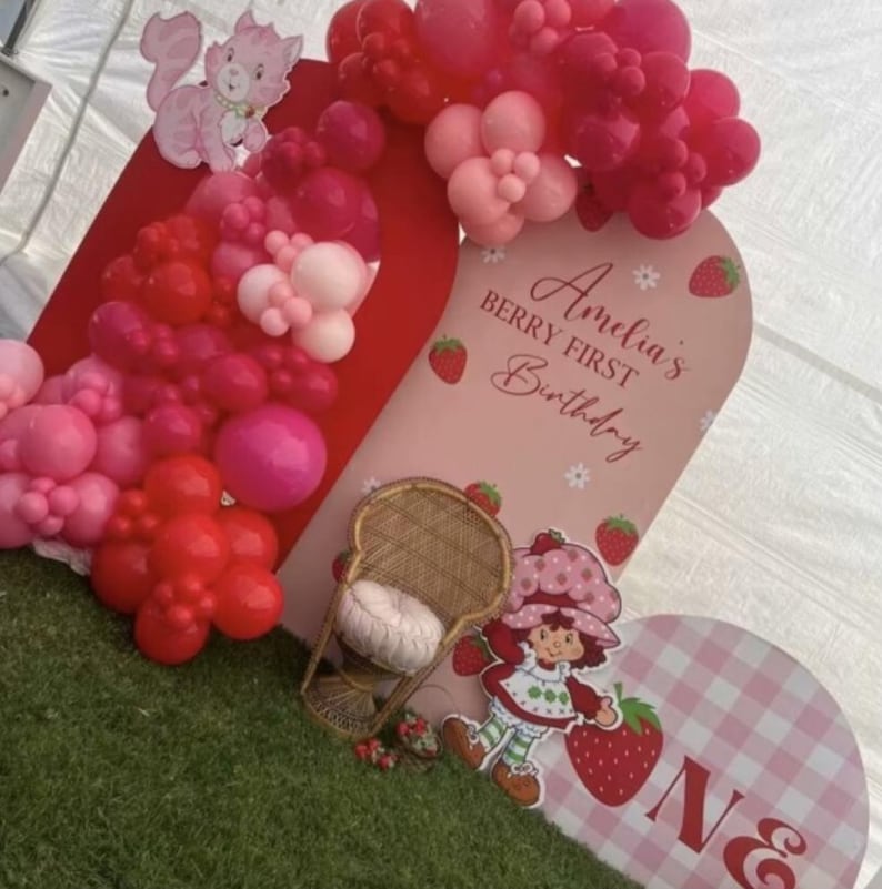 May include: A birthday display featuring a red and pink balloon arch, a pink arched backdrop with the text Amelia's BERRY FIRST Birthday, a wicker chair with a white cushion, a Strawberry Shortcake cutout, and a pink gingham sign with the letter ONE.