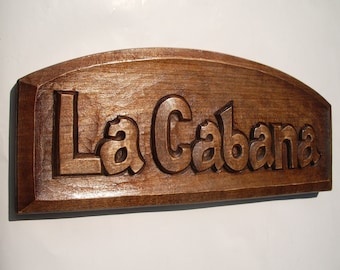Puede incluir: Un letrero de madera con el texto "La Cabana" tallado en él. El letrero está teñido de un color marrón oscuro y tiene una parte superior redondeada.