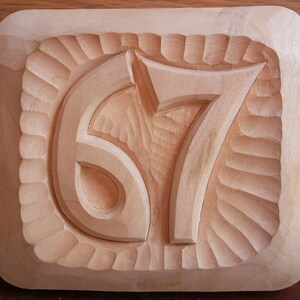 May include: A wooden plaque with the numbers 67 carved into it. The numbers are raised and have a textured surface. The plaque is a light brown color.