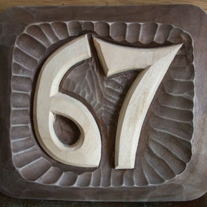 May include: A wooden plaque with the number 67 carved into it. The plaque is brown and the numbers are white.