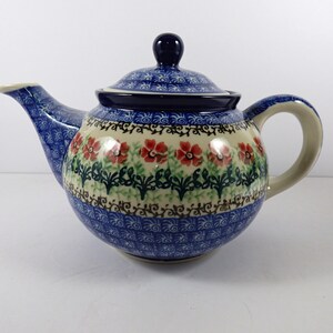 May include: A ceramic teapot featuring a cream-colored body decorated with a band of red flowers and green foliage. The lid, spout, and base are adorned with a blue and white patterned design. The teapot includes a matching lid with a knob.