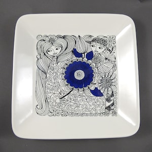 K&ouml;nnte beinhalten: Quadratischer wei&szlig;er Keramikteller mit einem schwarz-blauen Blumendesign. Das Design zeigt zwei stilisierte Figuren, eine gro&szlig;e blaue Blume und komplizierte schwarze Linienkunst. Der Teller hat abgerundete Ecken und eine gl&auml;nzende Oberfl&auml;che.