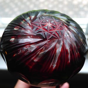 May include: A close-up of a ruby red glass object with a starburst pattern carved into the top. The glass has a deep red color and is highly reflective, with light reflecting off the carved facets.